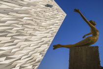 Ireland, Belfast, Titanic Quarter, Titanic Belfast Visitor Experience, 'Titanica' sculpture by Rowan Gillespie with a section of the building and blue sky in the background.