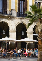 Spain, Catalonia, Barcelona, People sitting at tables under umbrellas for shade at a bar in the large historic square Placa Reial in the Gothic Quarter.