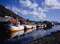 Norway, Lofoten, Napp, Line of traditional fishing boats moored beside harbour warehouses and reflected in flay calm water.