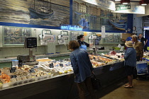 France, Pays Basque, St Jean de Luz covered market fish merchant. Customers inspecting fish, staff weighing out fish.